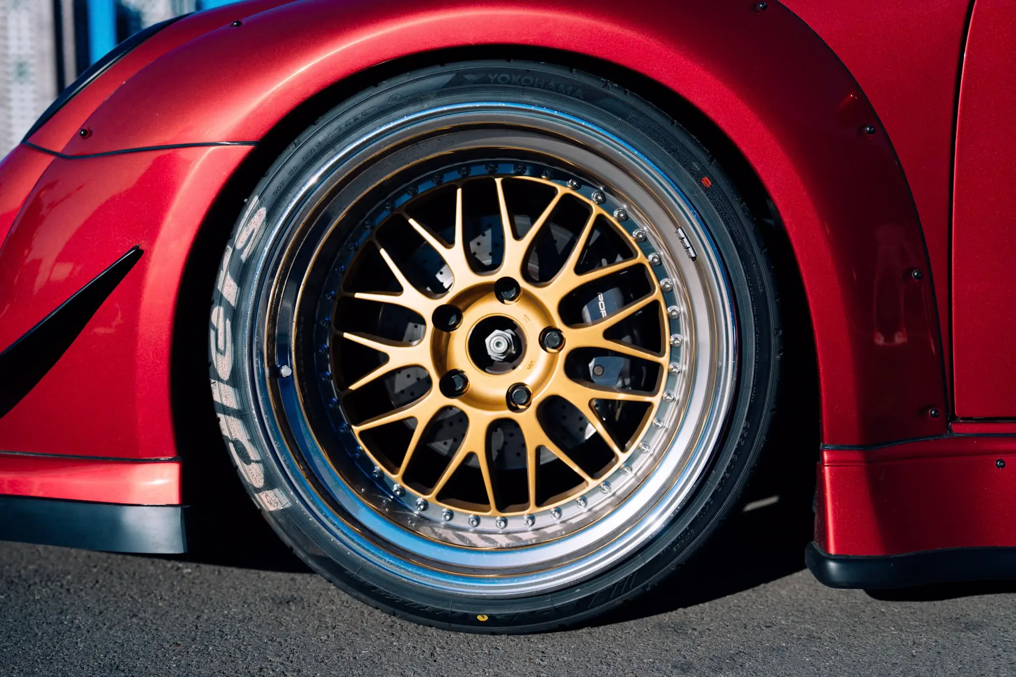Close-up of a gold multi-spoke alloy wheel with polished lip on a red car with wide body kit, sunlit street
