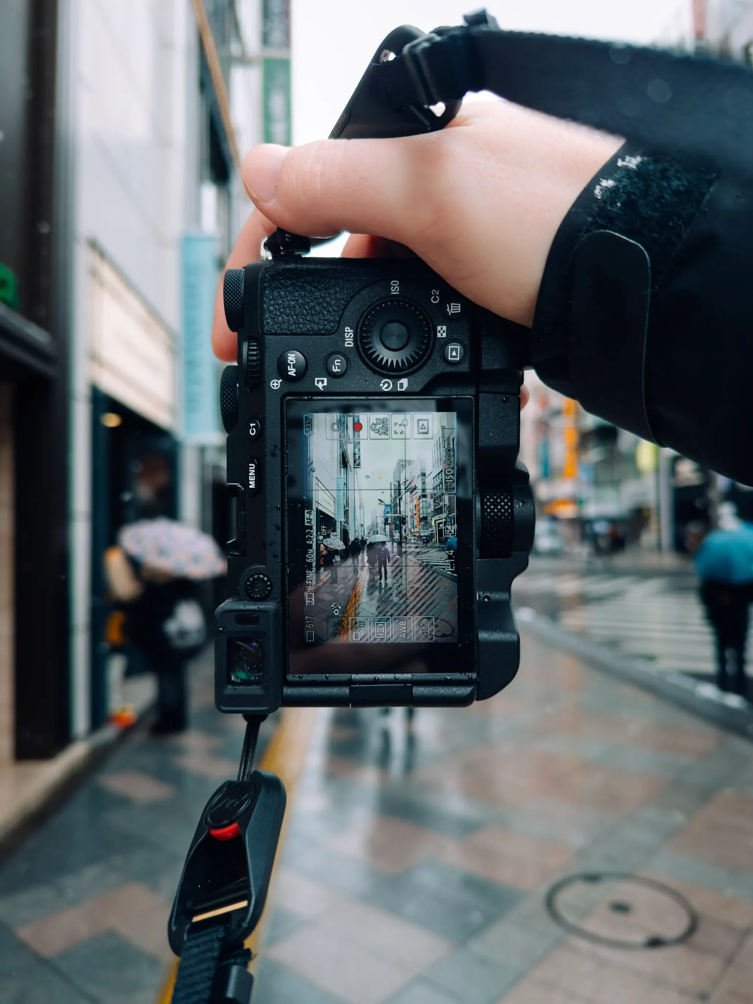 Camera held up showing a rainy city street with pedestrians and umbrellas on its LCD screen, blurred street behind