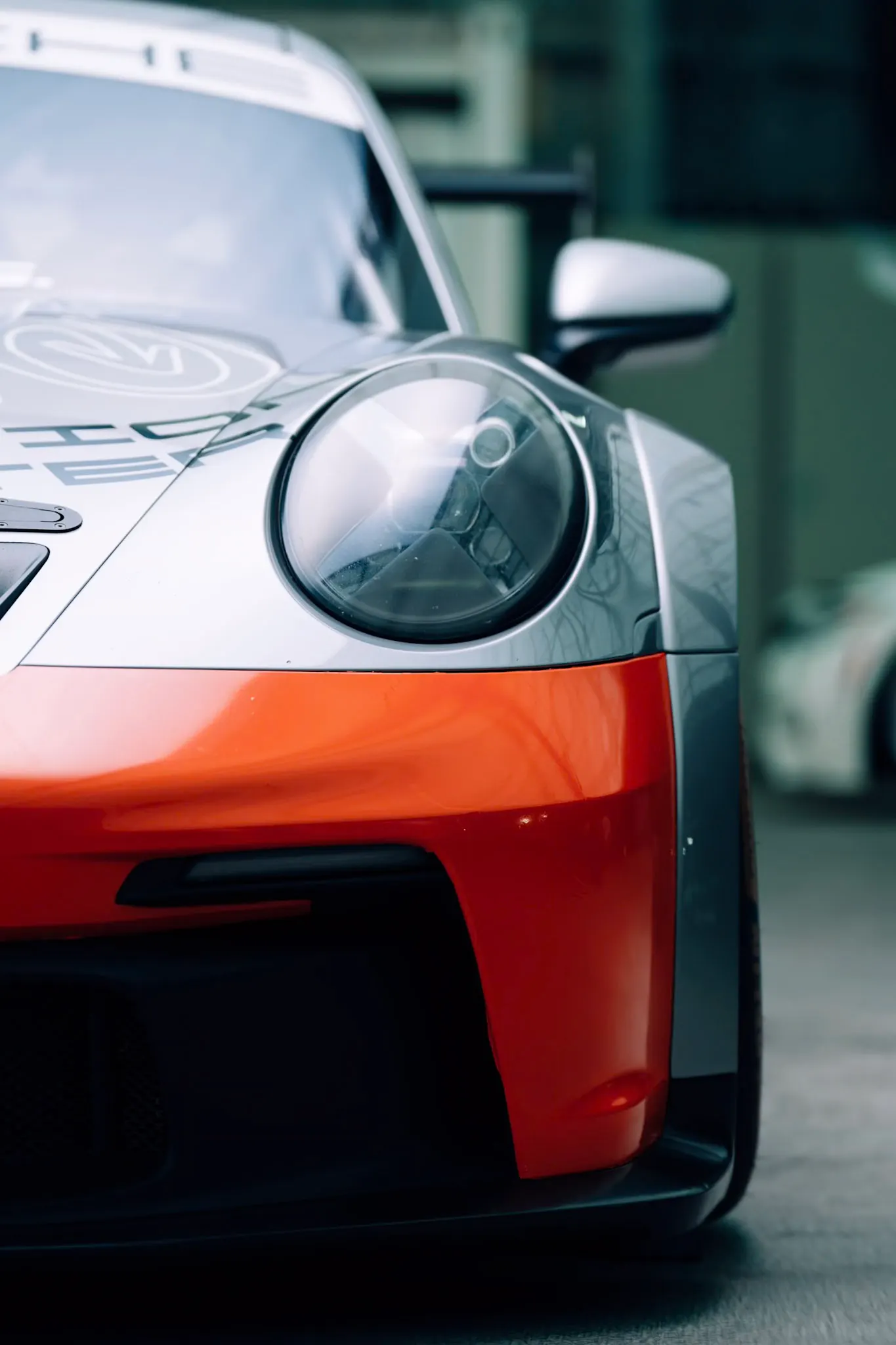Close-up of a silver and orange race car's front fascia and round headlight, with blurred background and soft natural light.