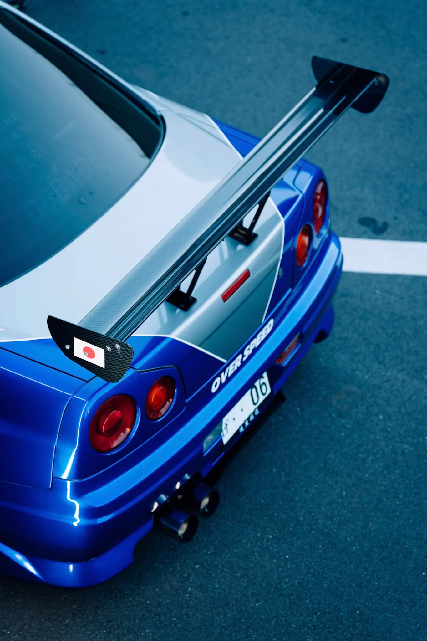 Rear view of a blue Nintendo Skyline GT-R sports car with a carbon fiber rear spoiler and Japanese flag decal on an asphalt lot