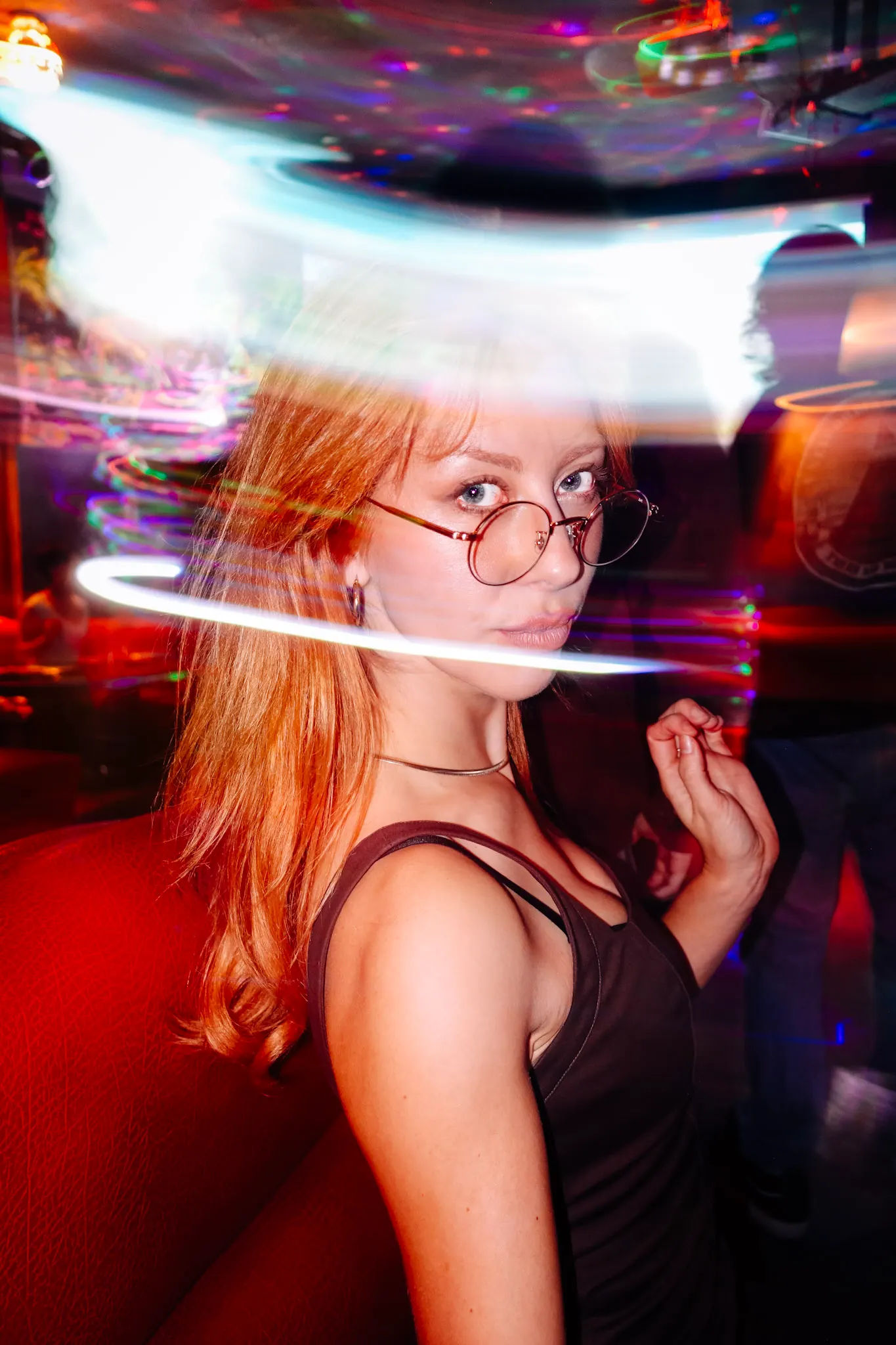 Woman with round wire-rimmed glasses and red hair in a dim bar, colorful light trails swirling behind her, red booth in foreground
