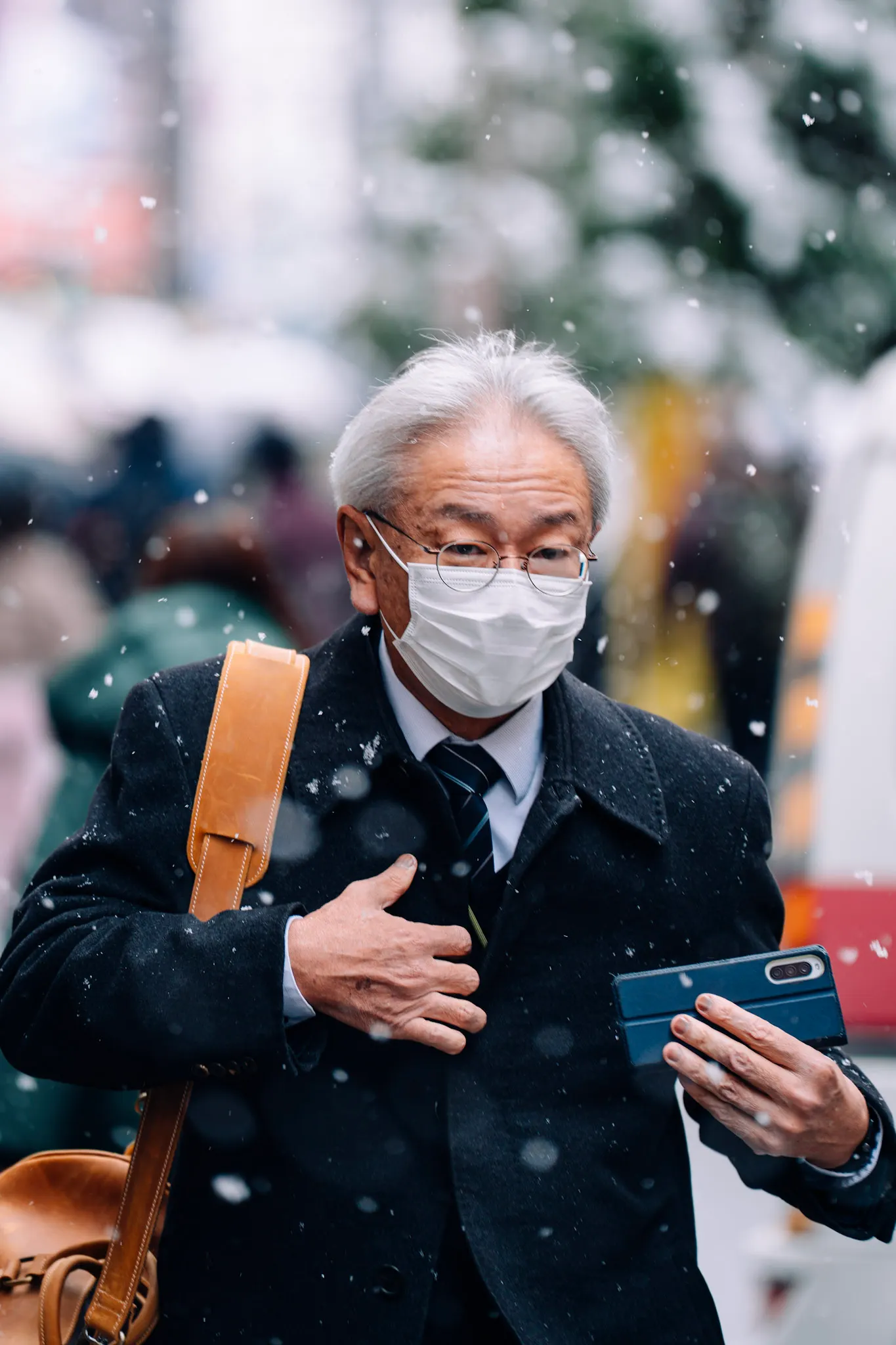 Older man in a face mask and glasses, hand on chest, holding a phone while snow falls on a city street