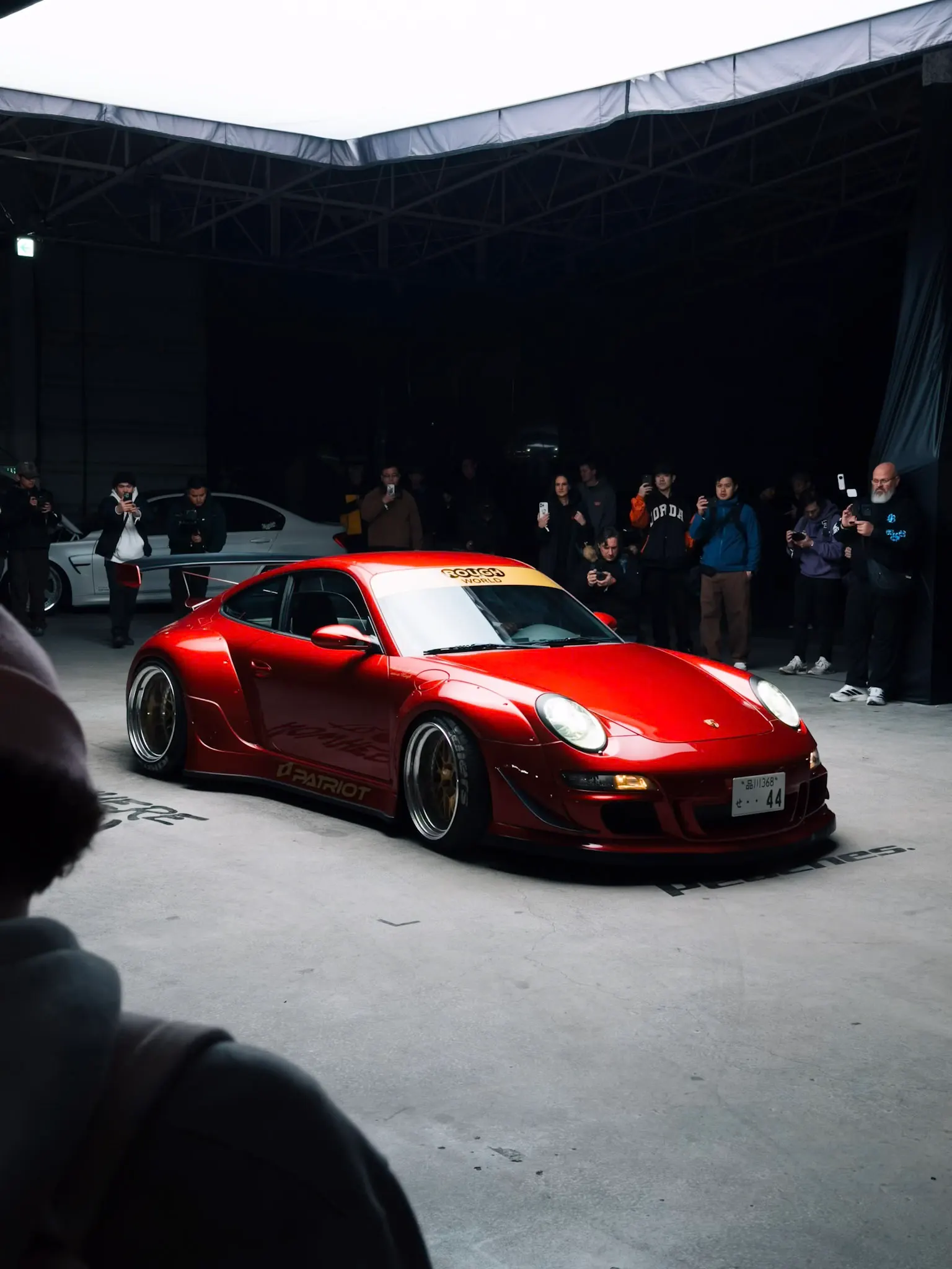 Red widebody Porsche 911 with aftermarket wheels displayed indoors while a crowd of spectators photographs it under dim lighting