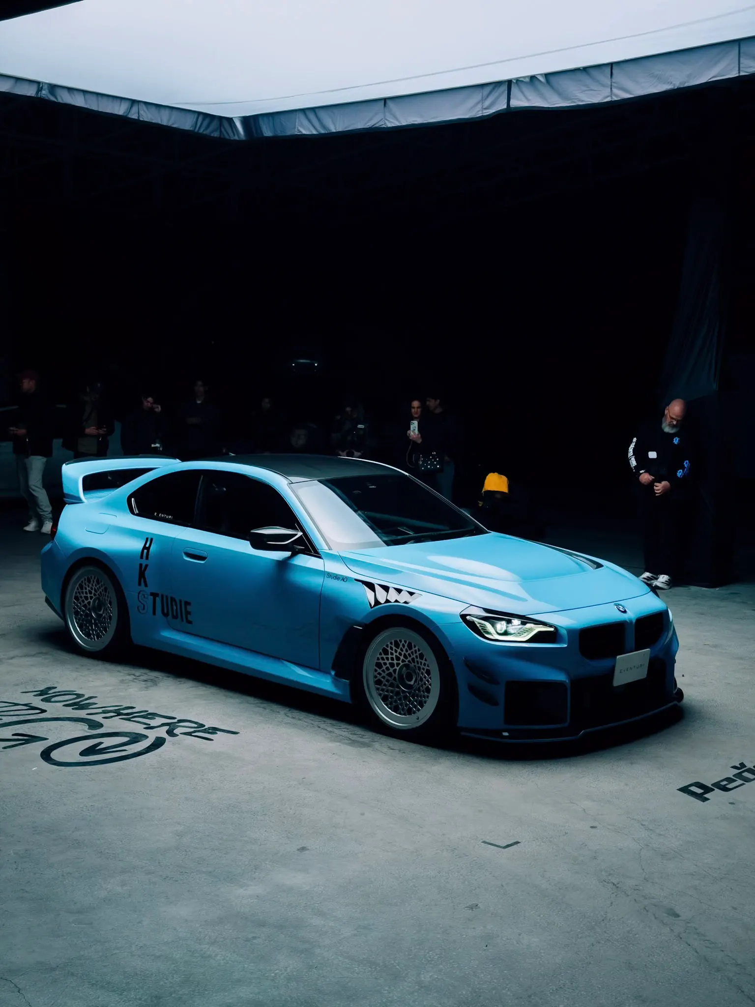 Matte blue BMW M2 with aftermarket wide body and mesh wheels displayed indoors under dark tent lighting, spectators in background