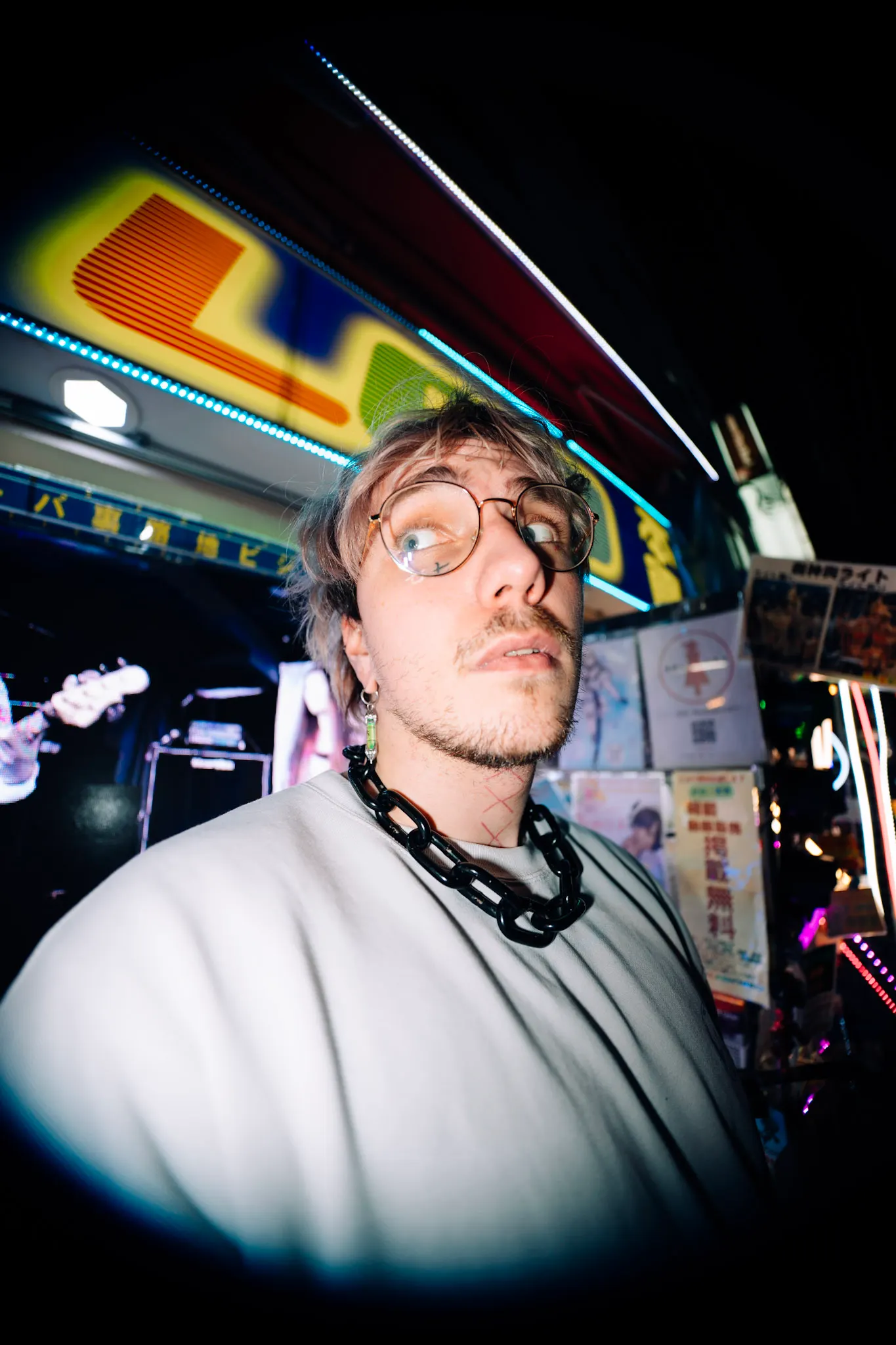 Man with round glasses and a chain necklace making a wide-eyed face under neon signs in a dim bar, with a guitar visible behind.