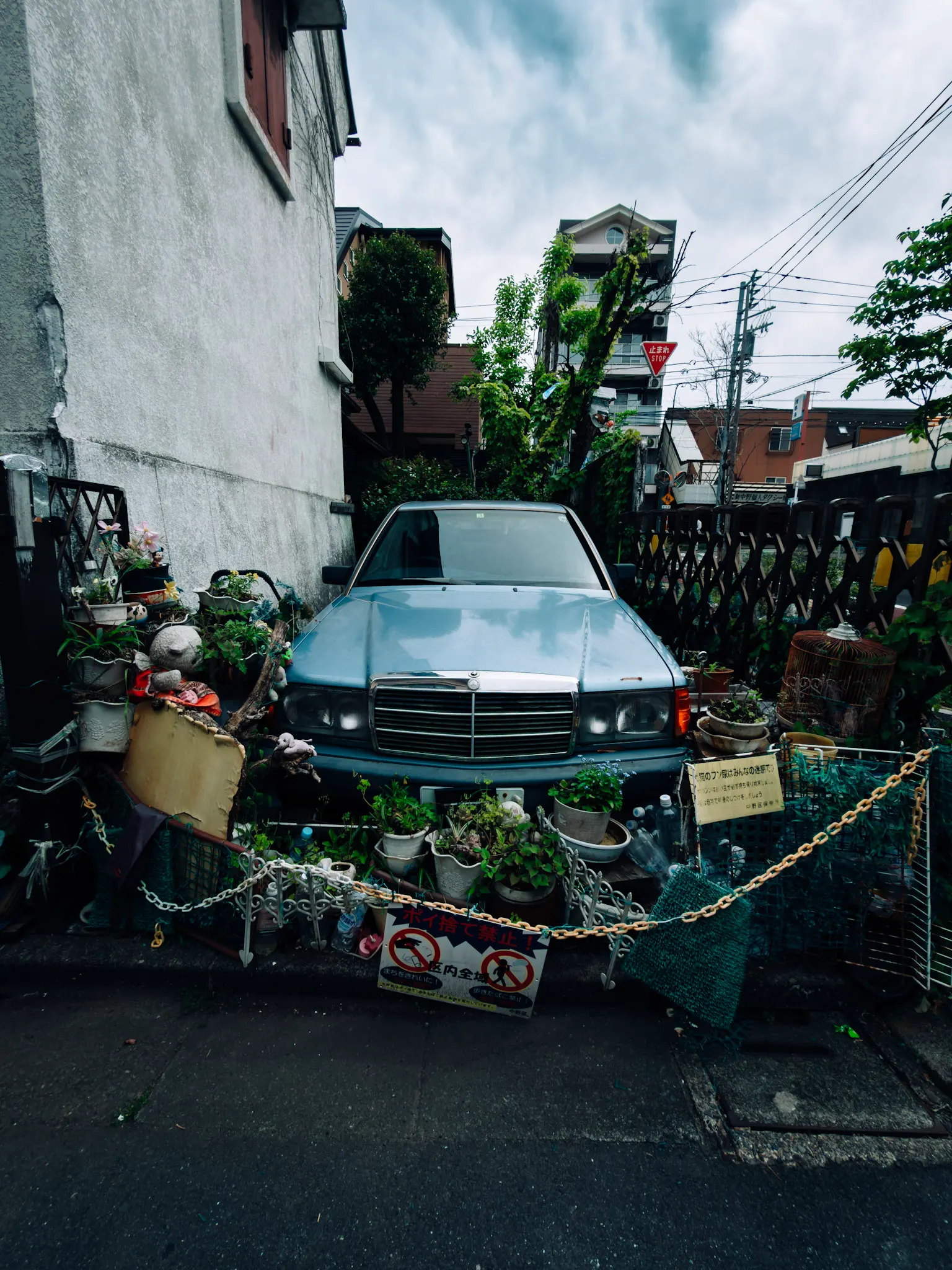 Light blue Mercedes sedan parked in a narrow alley, surrounded by potted plants, a chain barrier and cloudy sky