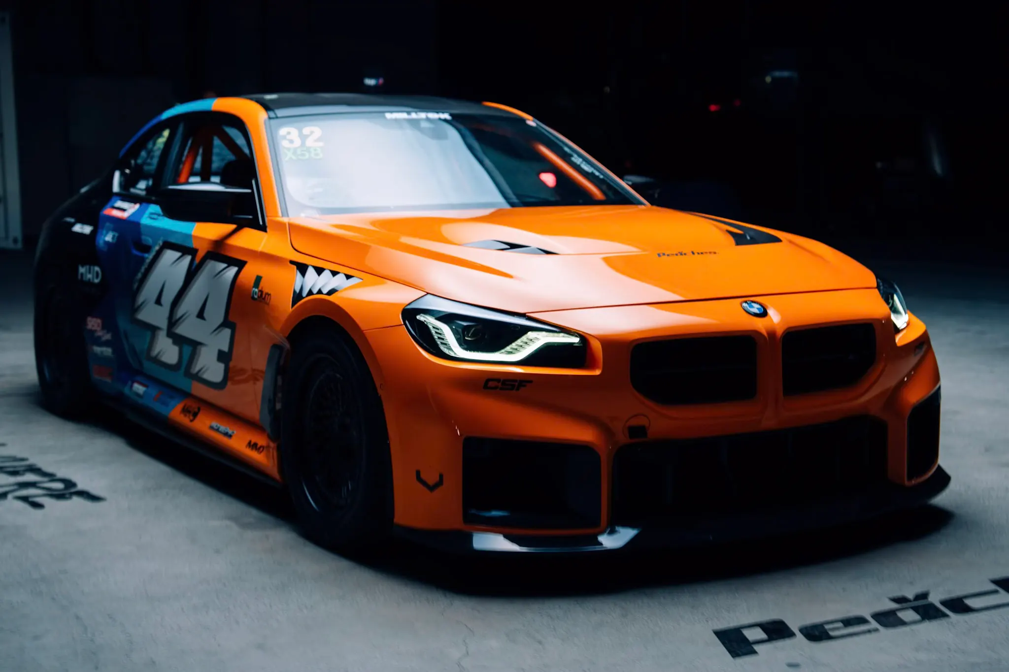 Orange BMW M2 race car with number 44 livery and checkered accents, lit dramatically against a dark concrete garage interior