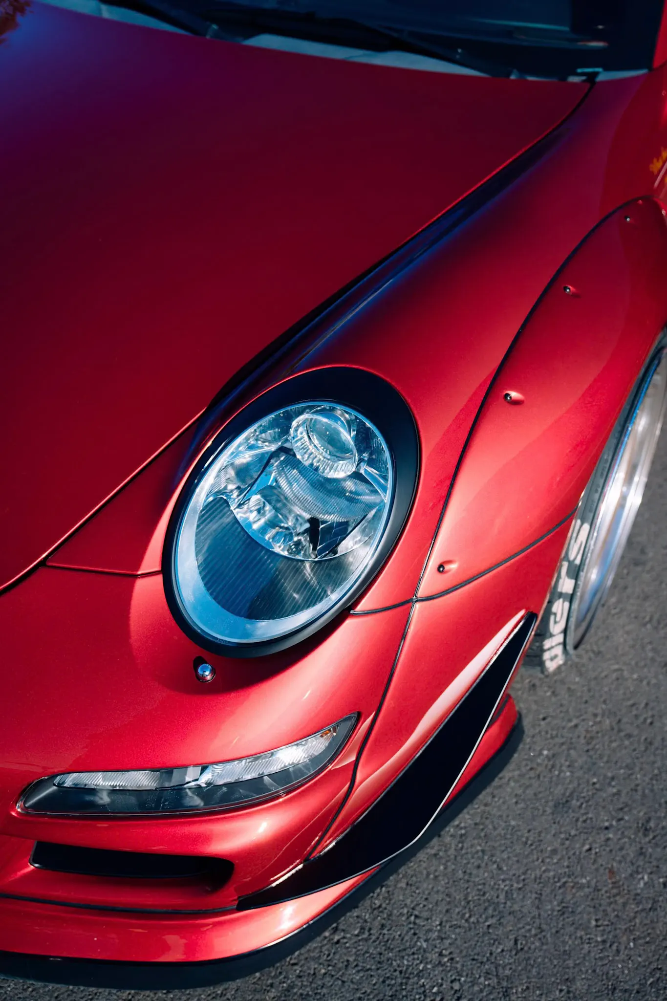 Close-up of a red modified sports car's round headlight, widened fender and carbon front splitter in warm daylight