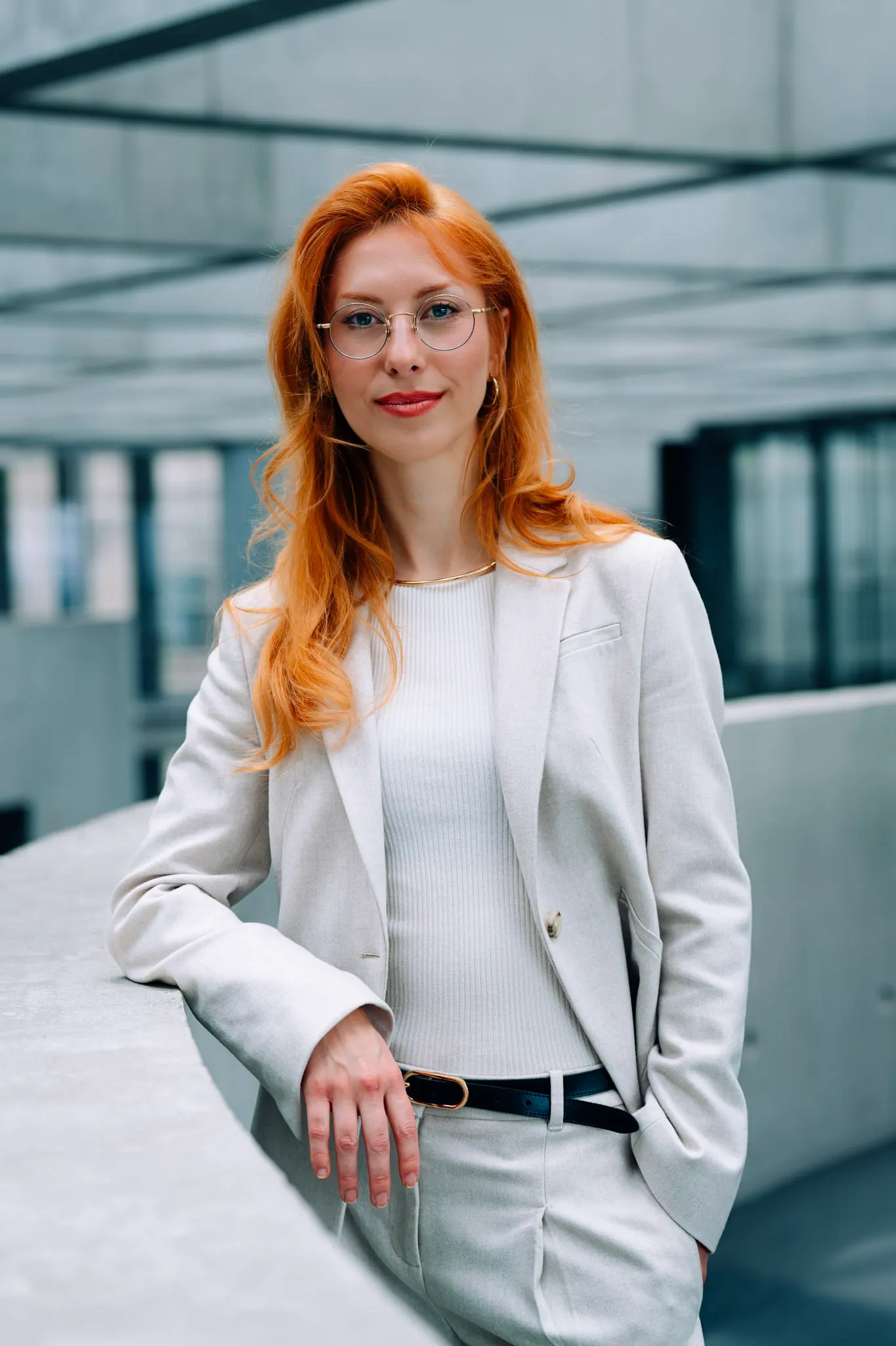 Woman with wavy red hair and round glasses in a light beige blazer, leaning on a concrete railing in a modern building.
