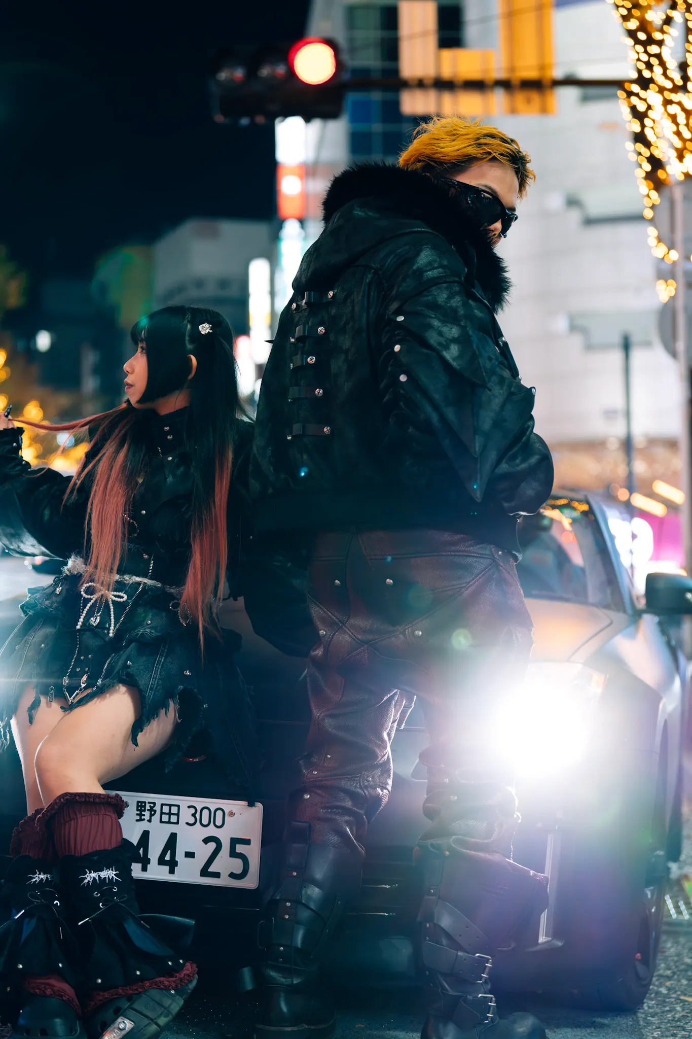 Two people in dark studded leather outfits leaning on a car at night, city lights and traffic signal glowing behind them